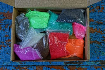  plastic bags with colored sand in an open paper box stands on a blue table