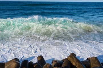 Blue ocean waves crash on a tetrapods breakwater forming white foam.
