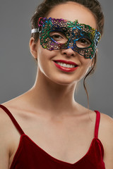 Portrait of smiling woman with tied back dark hair, wearing wine red crop top. The young girl is tilting head, wearing iridescent carnival mask with perforation. Vintage women's carnival accessory.