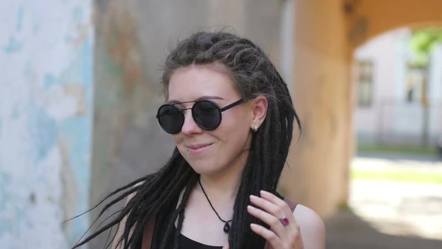 Portrait of an attractive girl with dreadlocks in sunglasses standing and smiling on a city street