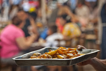 Roast potatoes in a large pan. Delicious food at the festival.A man pours a potato into a metal tray. Blurred people eat at the table in the background.