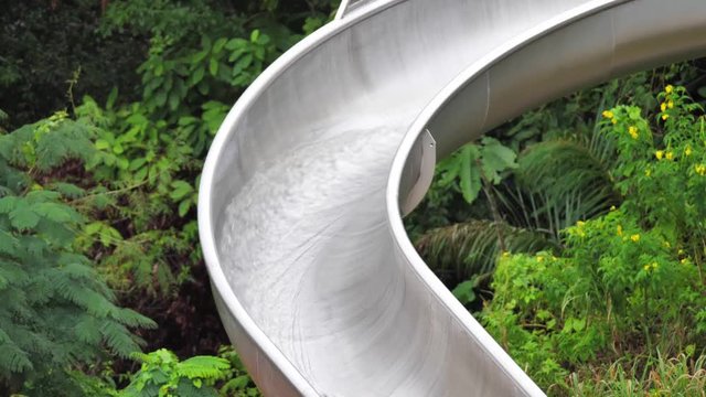 A closeup of water flowing and streaming down a curved half pipe open silver colored stainless steel water slide with lush green foliage and trees in the background.