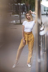 Obraz premium Gorgeous blond girl wearing yellow pants and white t-shirt, leaning on metal pipes near a modern building in the city. Street fashion concept. Modern fashion. 
