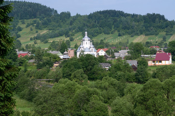 Fototapeta na wymiar View of the Carpathian village: catholic church, country houses, gardens and mountains. Lugi village, Ukraine