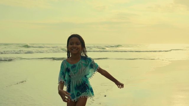 Gimbal steadycam tracking shot of 8 or 9 years old beautiful and happy Asian Indonesian child girl running carefree on the beach having fun enjoying Summer holidays on sunset at exotic island