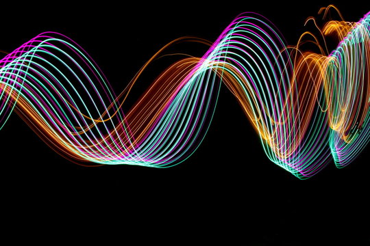 Long exposure photograph of neon purple, green and metallic gold colour in an abstract swirl pattern against a black background. Light painting photography.