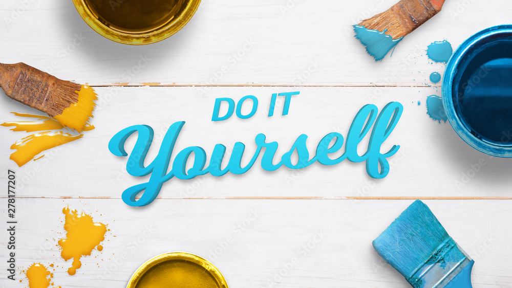 Poster do it yourself text on a white wooden table surrounded by color brushes, boxes and few color drops. 