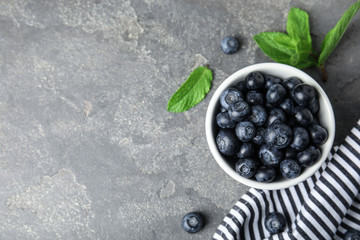 Bowl of tasty fresh blueberries, leaves and fabric on grey stone surface, flat lay. Space for text