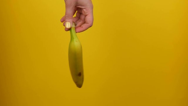 Female hand with yellow manicure holds a ripe banana fruit on a yellow background
