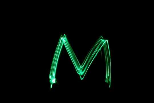 Long exposure photograph, light painting photography.  Letter m of an alphabet series, single letter, in neon green light, against a black background