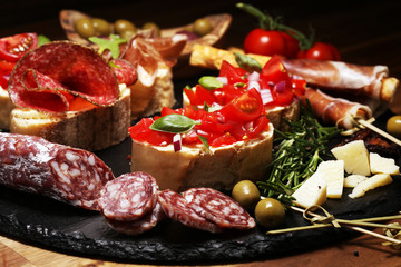 Traditional italian antipasto bruschetta appetizer with cherry tomatoes, cream cheese, basil leaves and balsamic vinegar on cutting board with prosciutto, salami, cheese