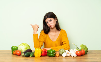 Teenager girl with many vegetables unhappy and pointing to the side