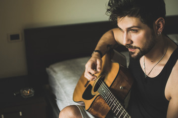 Italian Nude Boy playing Guitar sitting on bed smoking with light and shadows with uncombed hairs and beard