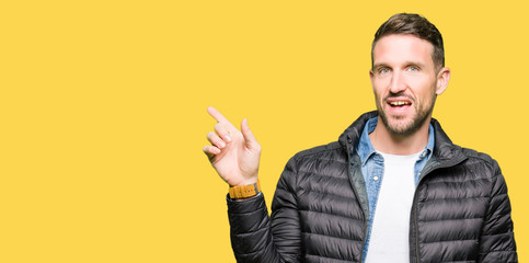 Handsome man wearing winter coat with a big smile on face, pointing with hand and finger to the side looking at the camera.