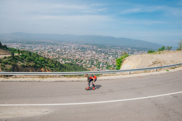 Male hipster wearing a hat and a red shirt is riding a longboard downhill on an open road