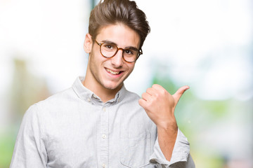 Young handsome man wearing glasses over isolated background smiling with happy face looking and pointing to the side with thumb up.