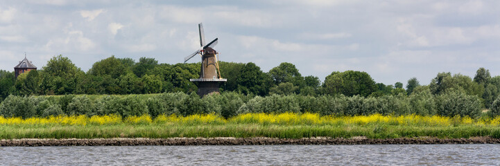 View on the shore of the Merwede River in the Netherlands, Typical Dutch landscape with traditional windmill to provide energy and power. Real Dutch weather with wind, clouds and sun.