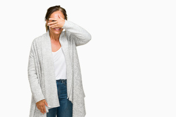 Beautiful middle age adult woman wearing winter sweater over isolated background peeking in shock covering face and eyes with hand, looking through fingers with embarrassed expression.