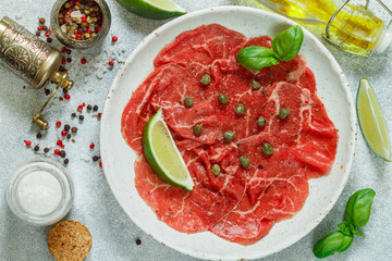 Delicatessen appetizer of fresh meat - beef carpaccio with lemon or lime, capers, olive oil, Basil, sea salt and freshly ground pepper on a white plate. Selective focus