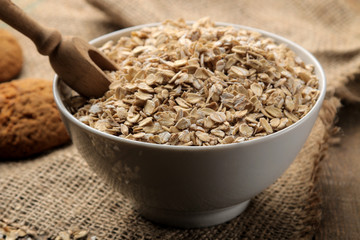 Dry oatmeal flakes in a white bowl and a wooden spoon on a brown wooden table