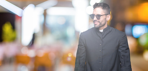 Young Christian priest wearing sunglasses over isolated background looking away to side with smile...