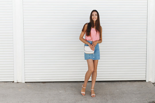 Young brunette woman wearing blue denim mini skirt, pink tank top, block heeled sandals, white cross body bag posing near white roller door. Trendy casual summer outfit. Everyday look. Street fashion.