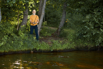 Fototapeta na wymiar Horizontal outdoor shot of serious healthy elderly man pensioner standing in bushes on river bank, holding angler or fishing rod cast in water. Angling, fishery and recreation. View from opposite side