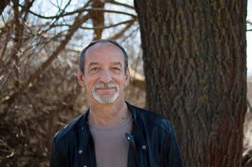 Male portrait of mature senior man with grey hair and beard in dark leather jacket standing outdoors near the tree on sky background in early spring.