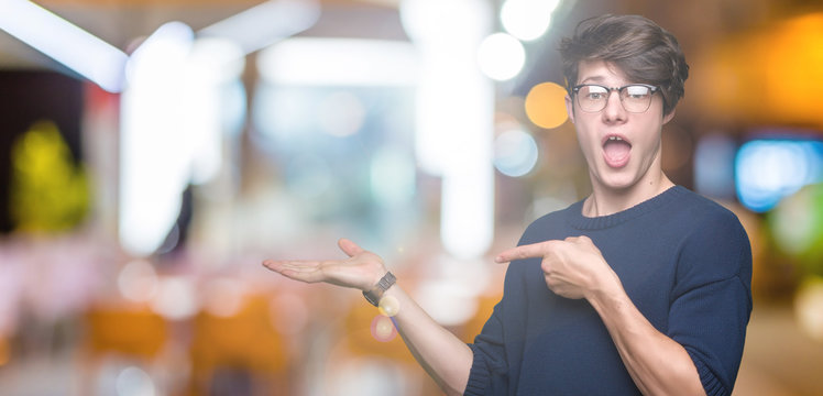 Young handsome man wearing glasses over isolated background amazed and smiling to the camera while presenting with hand and pointing with finger.