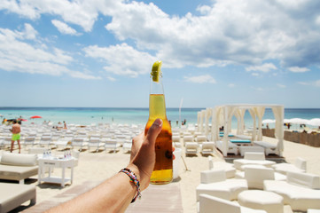 Male hand holding a bottle of beer against a sunny sky and crystal clear sea. In the background a beautiful beach resort. Vacation concept.