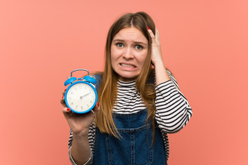 Young woman with overalls over pink wall restless because it has become late and holding vintage...