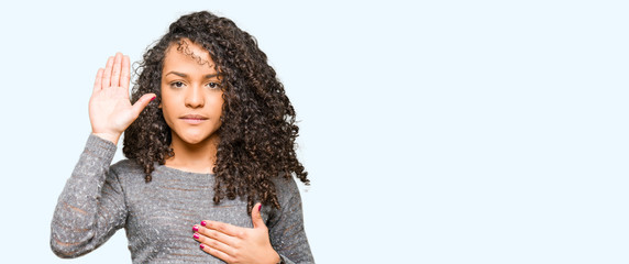 Young beautiful woman with curly hair wearing grey sweater Swearing with hand on chest and open...