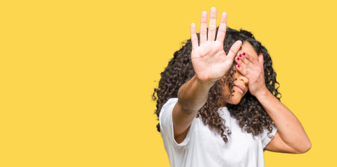 Young beautiful woman with curly hair wearing white t-shirt covering eyes with hands and doing stop...