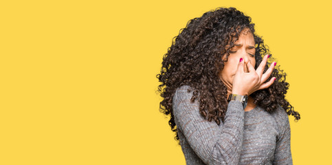 Young beautiful woman with curly hair wearing grey sweater smelling something stinky and...