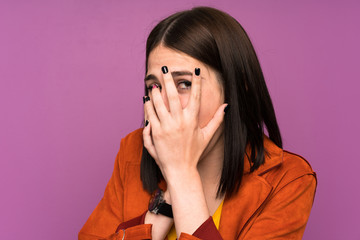 Young woman with a coat over isolated purple background covering eyes and looking through fingers