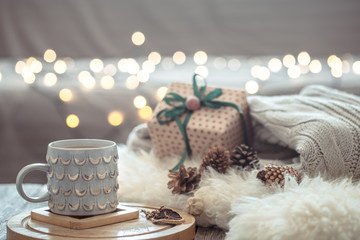 Obraz na płótnie Canvas Coffee cup over Christmas lights bokeh in home on wooden table with sweater on a background and present. Holiday decoration, magic Christmas