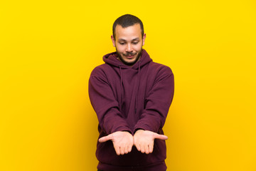 Colombian man with sweatshirt over yellow wall holding copyspace imaginary on the palm to insert an...