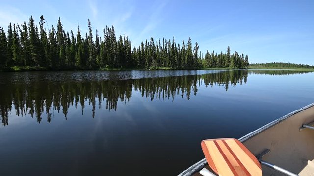 Canoeing along a calm northern river with trees and clouds reflecting in the water including a canoe and paddle in the foreground