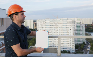 man with blank board in his hand in a protective helmet is standing on the roof of a building overlooking the buildings. Construction, architecture and business engineer at a construction site 