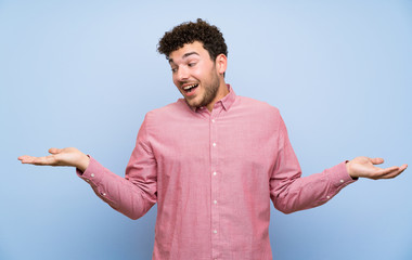 Man with curly hair over isolated blue wall holding copyspace with two hands