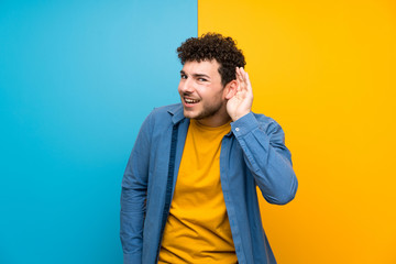 Man with curly hair over colorful wall listening to something by putting hand on the ear