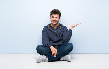 Young man sitting on the floor holding copyspace imaginary on the palm to insert an ad