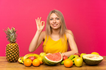 Young blonde woman with lots of fruits showing an ok sign with fingers