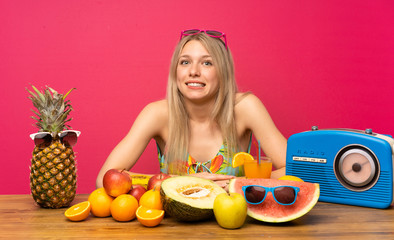 Young blonde woman with lots of fruits having doubts and with confuse face expression