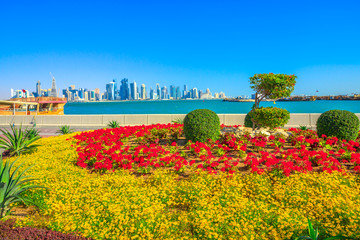 Obraz premium Spring landscape of flower beds and blooming trees on Corniche promenade of Doha Bay in Qatar. Tall skyscrapers of Doha Downtown skyline on distance. Blue sky in sunny day. Copy space.