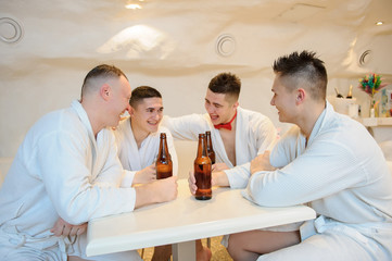 close up photo of 4 men in white gowns sitting around a table and drinking beer after sauna procedures