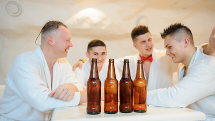 close up photo of 4 men in white gowns sitting around a table and drinking beer after sauna procedures