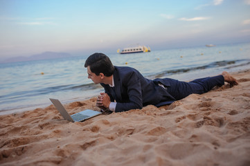 lateral view of a young man in suit with laptop lies on the beach working on the seaside