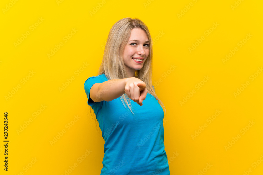 Wall mural young blonde woman over isolated yellow background points finger at you with a confident expression