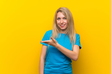 Young blonde woman over isolated yellow background presenting an idea while looking smiling towards